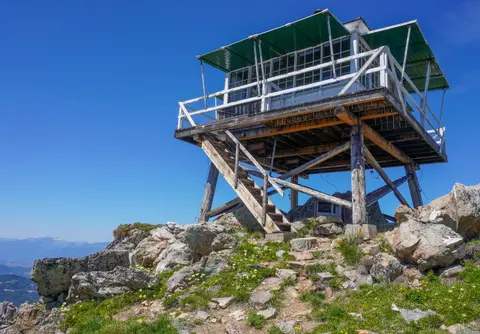 A fire lookout tower stands on a rocky mountain peak under a clear blue sky.