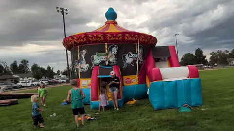 Children play near a large inflatable carousel bounce house with a slide on a grassy field.
