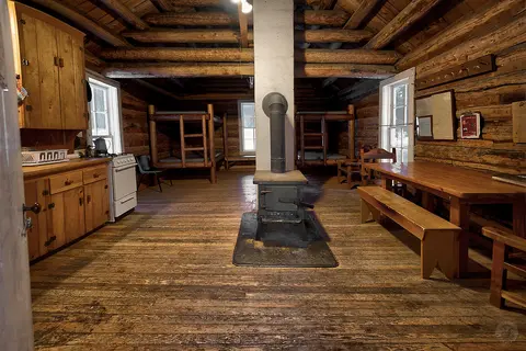 The interior of a rustic log cabin with bunk beds, a wood-burning stove, and a dining table.