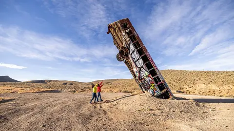 Two people pose next to a bus stuck vertically in the ground in the desert.