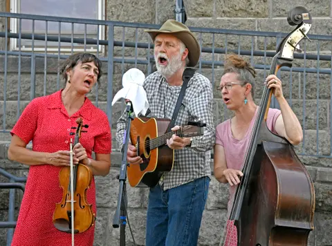 Three musicians sing and play instruments outdoors in front of a stone wall and metal railing.