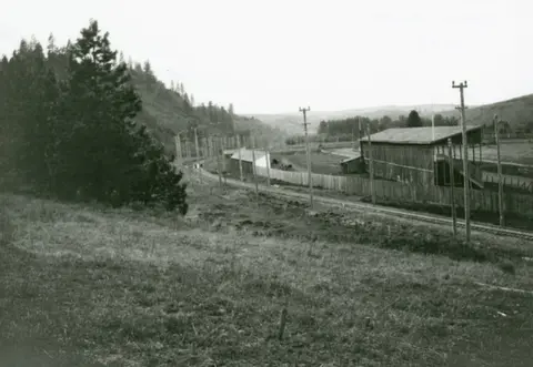 Historic black and white photo of a railway track running alongside a forested hill and wooden buildings.