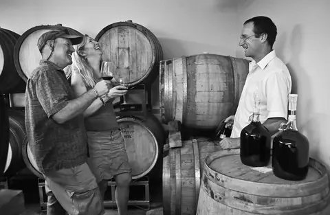 Black and white photograph showing three people laughing and holding glasses of wine inside a wine cellar with large wooden barrels.
