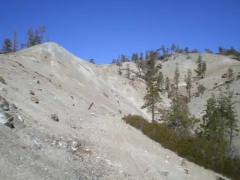 A landscape showing a dry, rocky hillside with sparse trees under a blue sky.