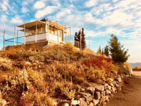 A fire lookout tower sits atop a hill covered in autumn foliage under a cloudy sky.