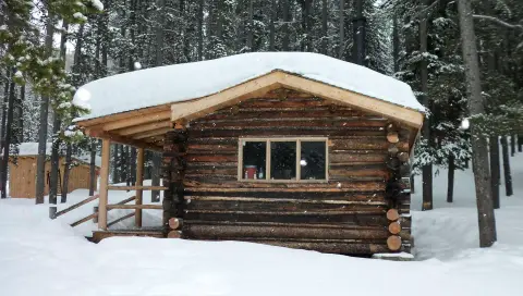 A rustic log cabin sits in a snowy forest surrounded by pine trees.