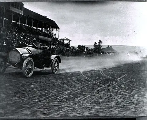 A historic photograph showing a vintage race car on a dirt track with spectators in the background.