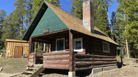 A rustic log cabin with a brick chimney is set in a wooded area.