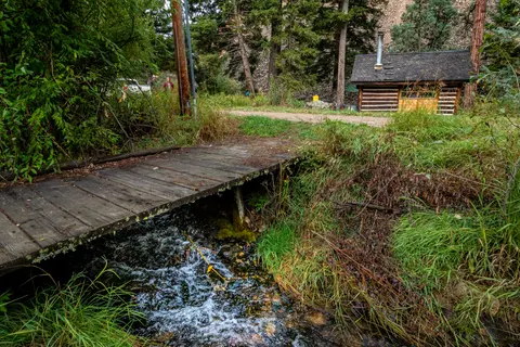 A small wooden bridge crosses a rushing stream with a cabin in the background.