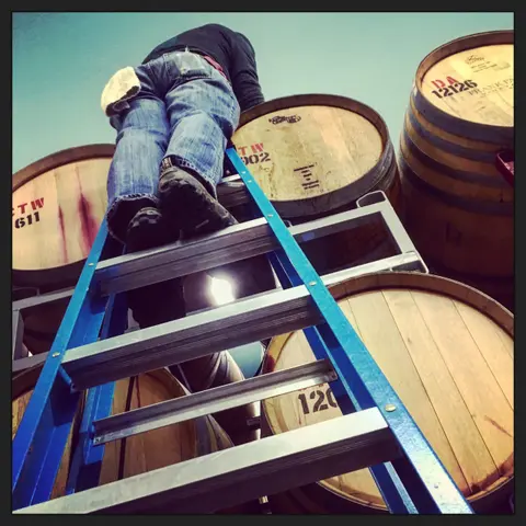 A person on a ladder reaches toward stacked wooden barrels.