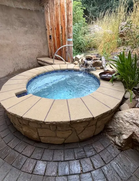A round stone hot tub is set outdoors with a small water feature and plants.