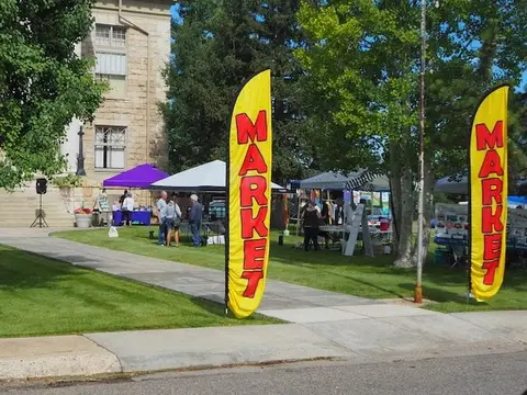 An outdoor market with yellow feather flags reading market set up on a lawn near a building.
