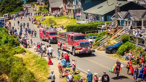 A parade with fire trucks travels down a street lined with spectators and coastal houses.