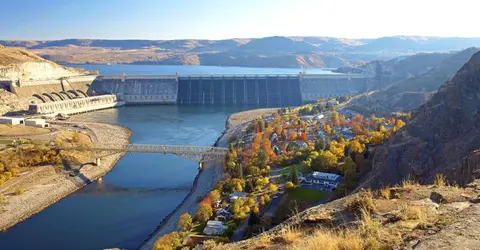 A large dam spans a river next to a bridge and a settlement surrounded by hills.