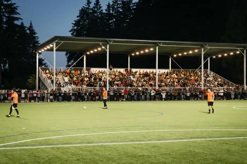 A soccer game is played at night in front of a packed covered grandstand.