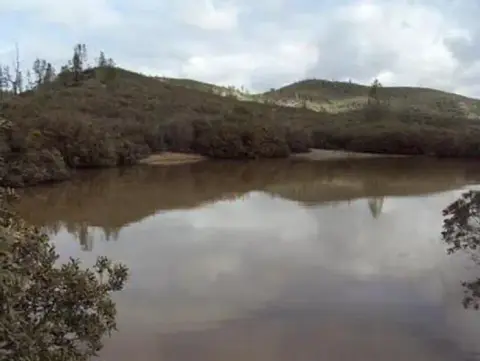 A still body of water reflects a cloudy sky surrounded by low hills and brush.