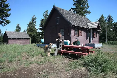 Two people and a dog by a cabin with a picnic table.
