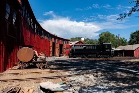 A train sits outside a red roundhouse under a blue sky.