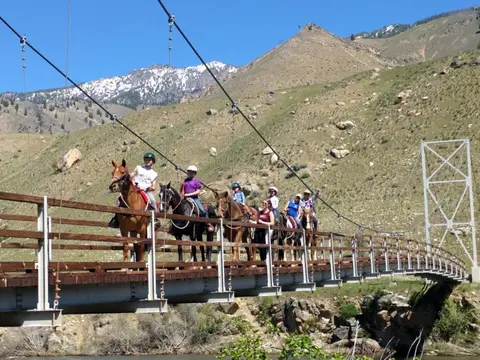 A group of people ride horses across a suspension bridge in a mountainous landscape.