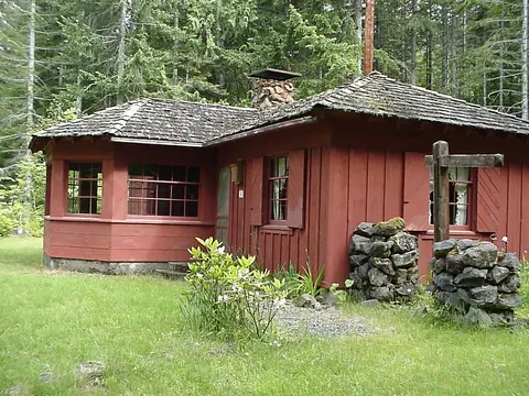 A red wooden cabin sits in a clearing surrounded by a dense forest.