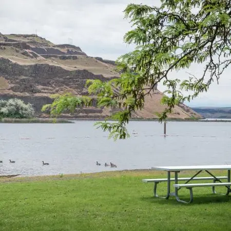 A picnic table on the grass near a body of water with hills in the background.