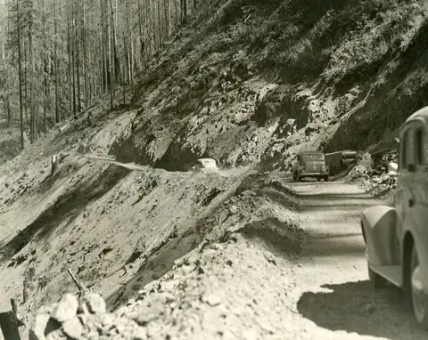 A historic black and white photograph showing vintage cars driving on a narrow dirt mountain road.