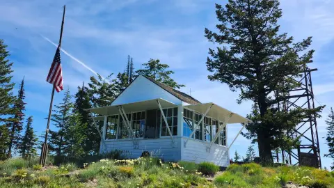 A small white building with a flagpole and an American flag stands outside among trees under a blue sky.
