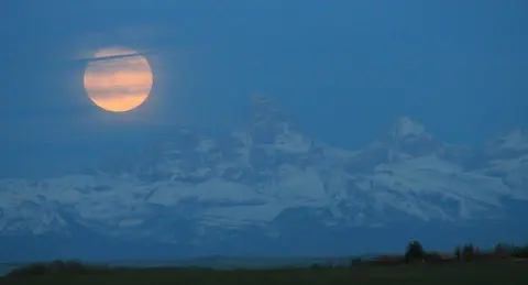 A full moon rises over a snowy mountain range.