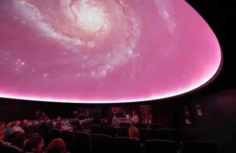 An audience seated inside a planetarium with a spiral galaxy projected on the dome ceiling.
