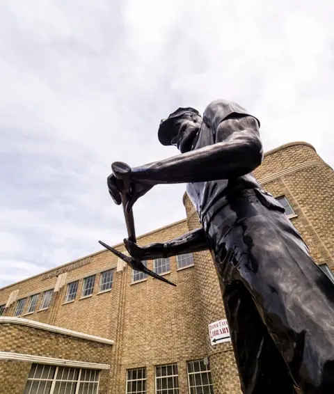 A low-angle shot of a dark metallic statue of a worker holding tools against a brick building and cloudy sky.