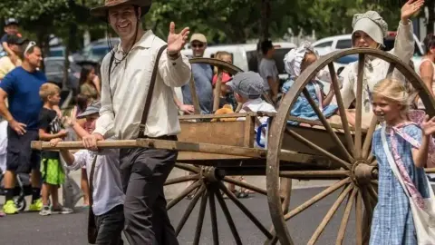 People in historical costumes wave while pulling a wooden cart in a parade.
