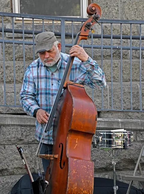 A street musician plays a double bass in front of a stone wall and metal railing.