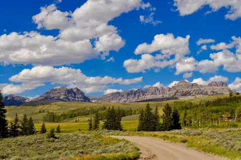 A dirt road winds through a green valley toward distant mountains under a blue sky with fluffy white clouds.