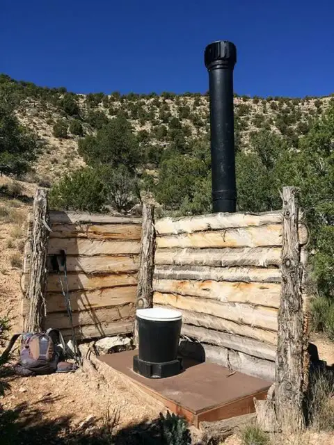 An outdoor toilet structure with wooden walls and a black ventilation pipe in a natural landscape.