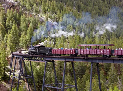 A steam train crosses a high trestle bridge through a forested mountain landscape.