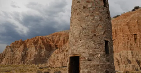 A stone tower stands in front of red rock formations under a cloudy sky.