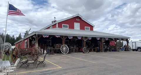 An exterior view of a red barn-like building displaying antique items and wagon wheels for sale outside.