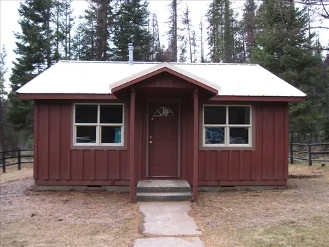 A small red cabin with a white metal roof stands in a wooded area.