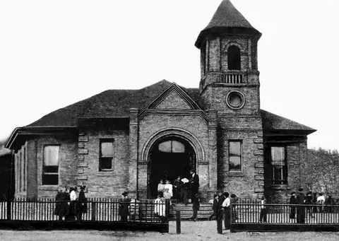 Black and white historic photograph of people gathered outside a brick building with a bell tower.