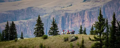 Three people horseback riding across a landscape with cliffs in the background.