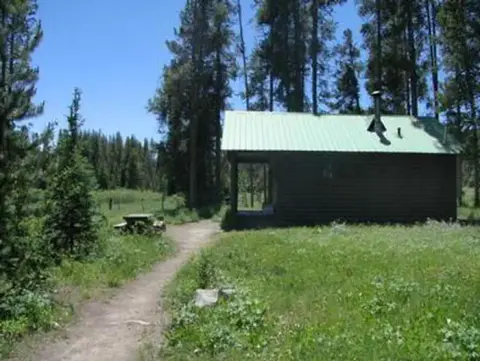 A small cabin sits in a clearing surrounded by trees and a dirt path.