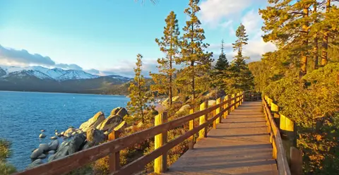 A wooden boardwalk runs alongside a blue lake with snowy mountains in the background.