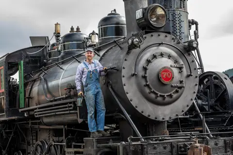 A man in overalls stands on the front of a steam locomotive.