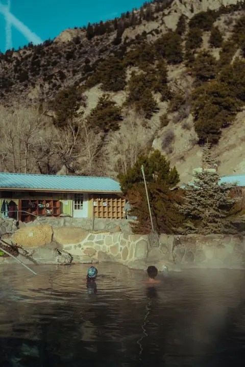 People soak in an outdoor hot spring pool in front of a mountain backdrop.