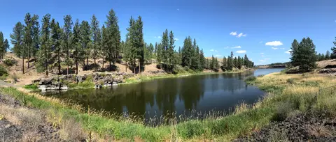 A wide panoramic view of a calm river bordered by trees and grassy banks under a blue sky.