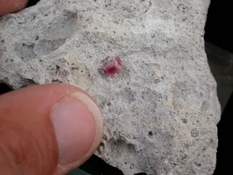 A close-up photo shows a small pink crystal embedded in a grey rock held by a person's fingers.