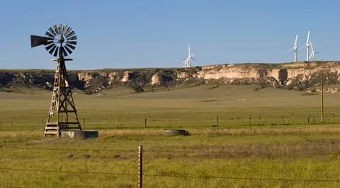 A traditional wooden windmill stands in a grassy field with cliffs and modern wind turbines in the background.