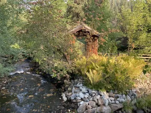 A small creek flows past lush green trees and a rustic wooden structure.