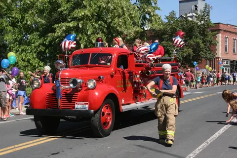A bright red vintage fire engine participates in a parade on a sunny street.