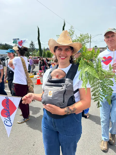 A woman wearing a cowboy hat and carrying a baby in a front pack attends an outdoor event.